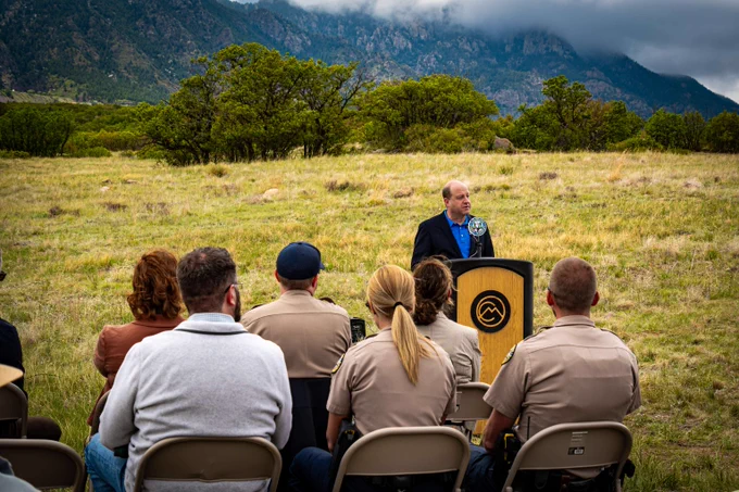 Governor Jared Polis speaks at a podium outdoors to an audience seated in folding chairs with mountains and a cloudy sky in the background.