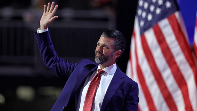 Donald Trump Jr., a white man, wears a suit and waves while standing in front of an American flag.