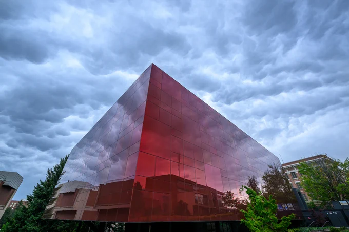 The crimson glass W S U Museum of Art has gray puffy clouds above. Several buildings are reflected in the glass. 