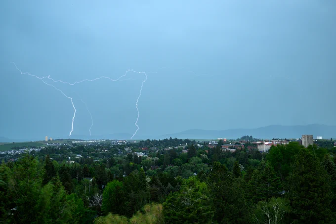 Color outdoor photo of lightning hitting outside of the City of Pullman. Gray skies are above green trees and various city buildings. 