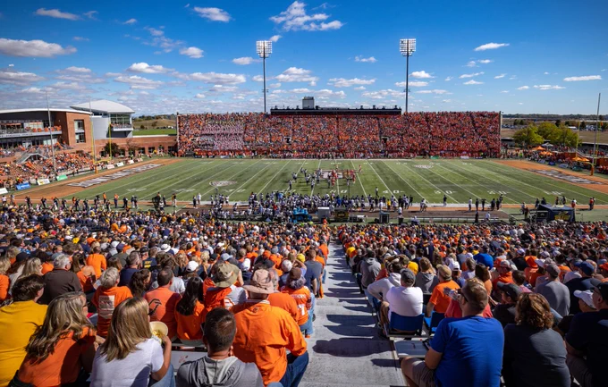 The image shows a crowded football stadium during a bright day. The field is lined with players, and the stands are filled with fans wearing orange and white. The sky is clear with scattered clouds.