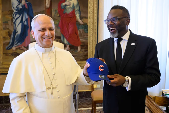 Mayor Brandon Johnson, looking to the camera in mid-speech, presents His Holiness Pope Leo XIV with a Chicago Cubs baseball cap, while the Pope reaches for the hat laughing. 