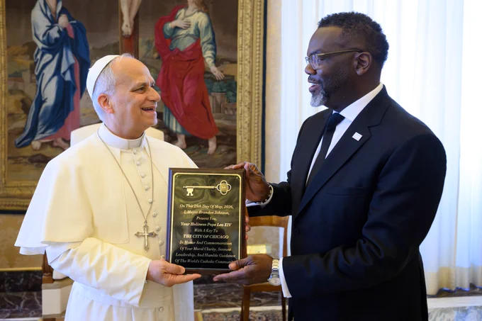 His Holiness Pope Leo XIV and Mayor Brandon Johnson look at one another laughing while Mayor Johnson hands the Pope the Key to the City of Chicago. The key placard reads, "On this 28th Day of May, 2026, I, Mayor Brandon Johnson, present to you, Your Holiness Pope Leo XIV, with a key ot the City of Chicago, in honor and commemoration of your moral clarity, servant leadership, and humble guidance of the world's Catholic community.