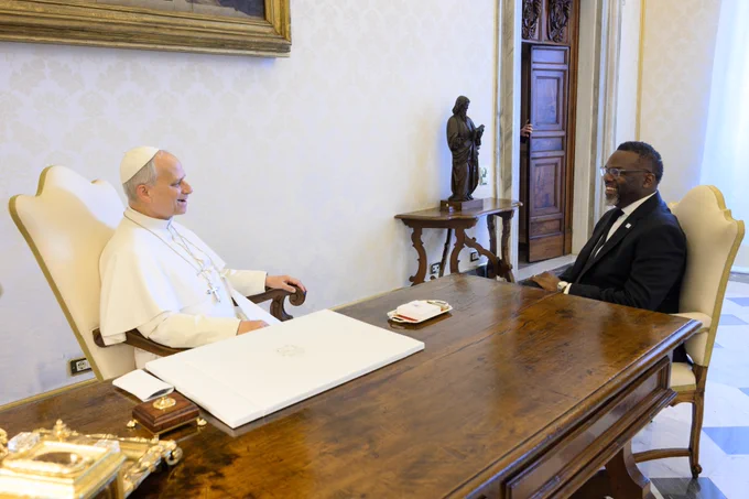 His Holiness, Pope Leo XIV, and Mayor Brandon Johnson are seated near one another at a wooden desk, smiling in discussion, with a statue of a saint against the wall behind and between them. 