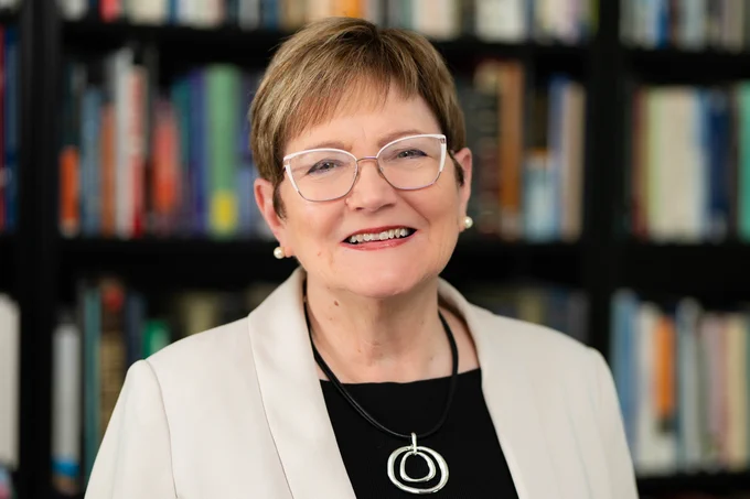 Ann Austin smiles in front of a bookshelf. Ann wears a black blouse, a beige blazer and a black necklace with a silver pendant. Ann's blonde hair is short. Ann has pearl earrings and silver-rimmed glasses.