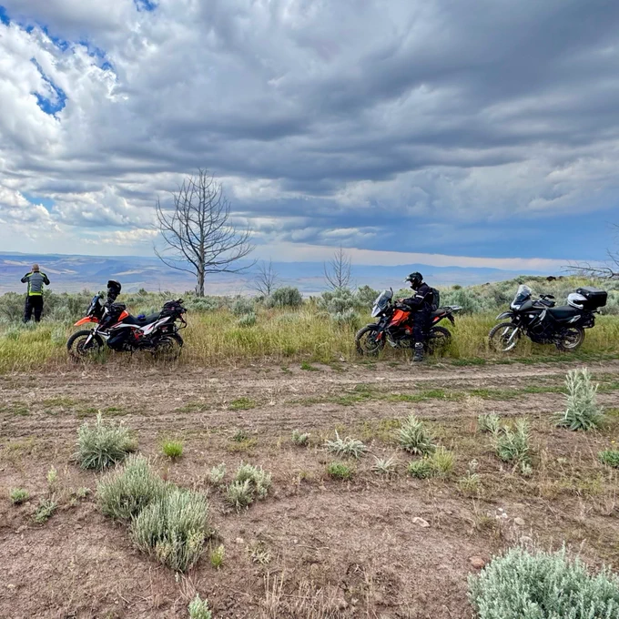 Three adventure motorcycles parked at the side of a forest road with a storm in the distance.