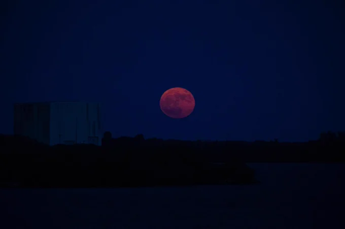  Una fotografía de una superluna azul sobre el Centro Espacial Kennedy en Florida durante la noche. La Luna se ve grande y rojiza sobre el horizonte, con siluetas oscuras de edificios, árboles y agua en primer plano.