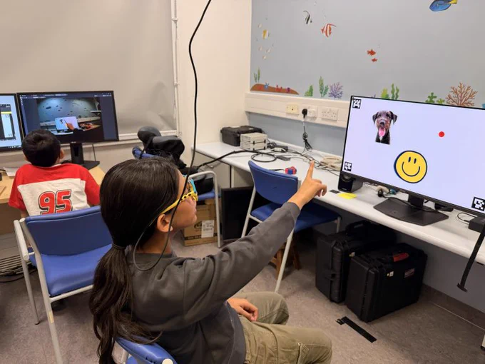 A child wearing eye-tracking glasses points at a computer screen displaying a dog image and a smiley face, while another young person sits at a nearby workstation in a lab-style room with multiple computers