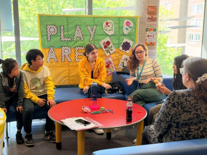 A small group of children and researcher Alicia Fothergill sit on blue sofas around a red table in a bright room, discussing and handling colourful objects. Behind them a colourful “PLAY AREA” poster is displayed on the wall.