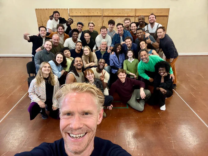 A selfie. A man holds the phone up and takes a photo of himself and a group of people gathered together in a rehearsal room.