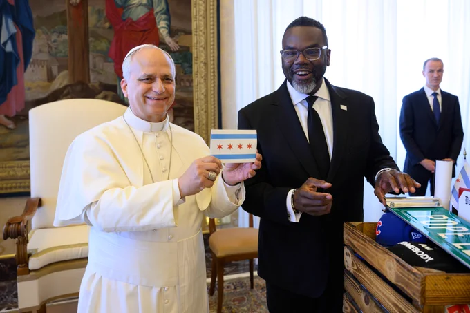 His Holiness, Pope Leo XIV holds up a card adorned with the Chicago Flag, smiling, while standing next to Mayor Brandon Johnson who is reaching into a basket of gifts from the Chicago delegation, including an honorary street sign bearing the Pope's name. 