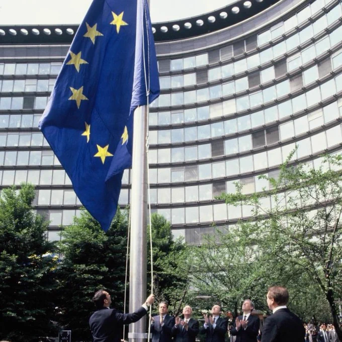 Hoisting of the European flag in presence of Charles Rutten, Jacques Delors, Pierre Pflimlin and Carlo Ripa di Meana. A person is raising a large European Union flag on a flagpole in front of a glass building (Berlaymont, Brussels). The scene is set outdoors with greenery and trees visible around.  
