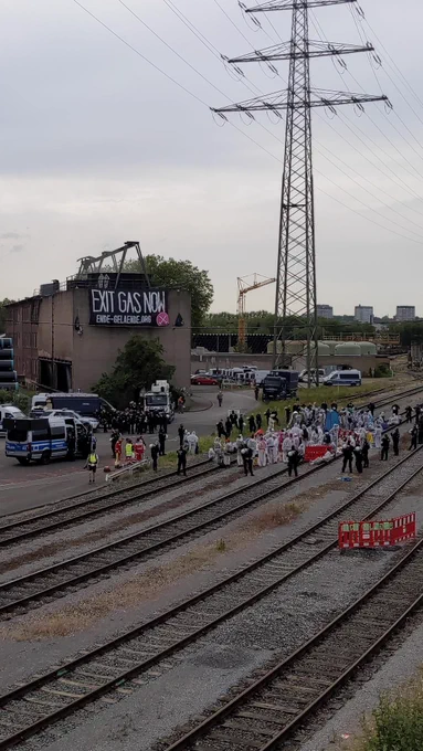 Im Vordergrund sind Bahngleise. Auf dem Bahngleisen sitzen 200 Aktivist*innen in weißen Anzügen. Diese sind von der Polizei umstellt. Einige Polizeiautos stehen neben den Schinen.
Hinter den Schienen liegen Pipelinerohre und eine Fertigungshalle mit einen Transparent ist zu sehen.  Auf dem Transparent steht 'Exit Gas Now' und ende-gelaende.org
Am Horizont befinden sich Kühltürme
