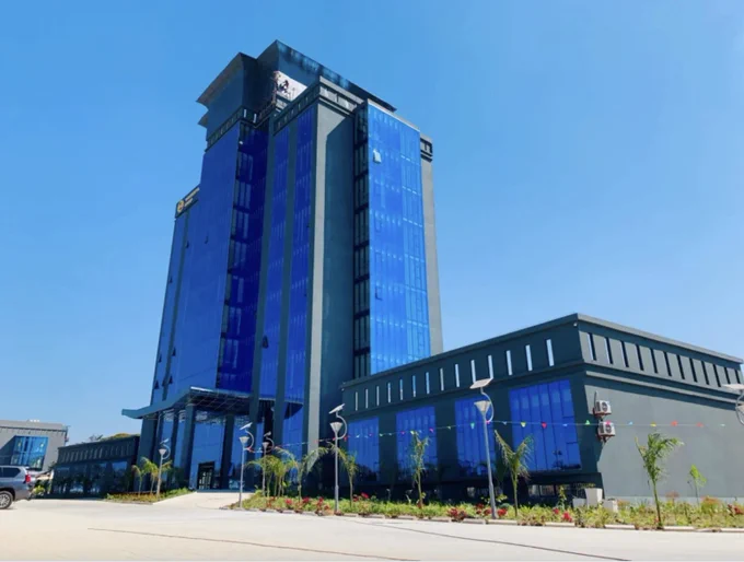 A tall, modern building with reflective blue glass windows and a smaller adjacent structure. The environment includes a clear sky, landscaped greenery, and a paved entrance area.