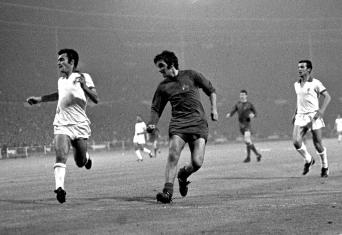 Black and white photo of George Best in dark uniform dribbling the ball past two opponents in white jerseys during a night match.