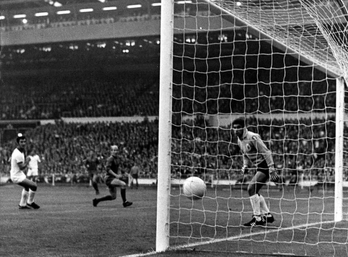 A light football crossing the goalline as Bobby Charlton and the Benfica goalkeeper watch closely during a crowded match.