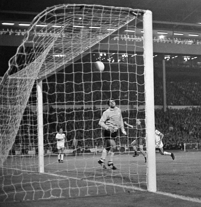 A light football flying into the net as the Benfica goalkeeper and Bobby Charlton in the background look on during a night match in a packed stadium.