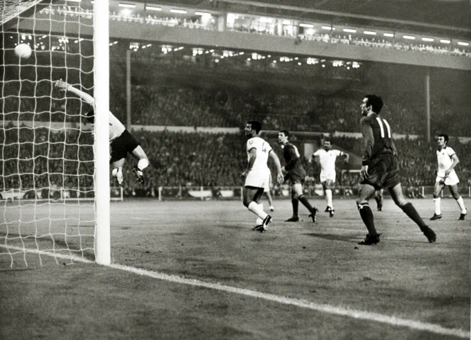 The Benfica goalkeeper leaps to try and save a ball heading toward the top corner of the goal during a night match in a packed stadium.