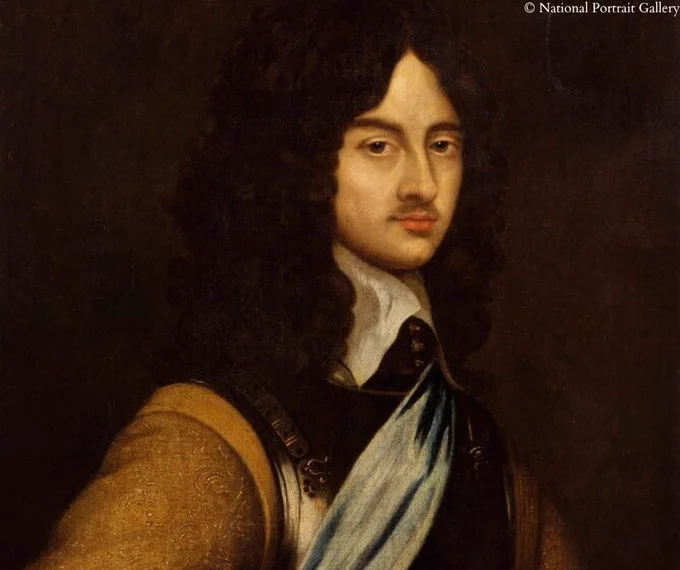A 17th-century portrait of King Charles II with long, dark, curly hair. He has a subtle mustache and is wearing an ornate brown coat with intricate patterns, a white shirt, and a dark vest. A light blue sash drapes across his chest. The background is dark, emphasizing his composed expression.