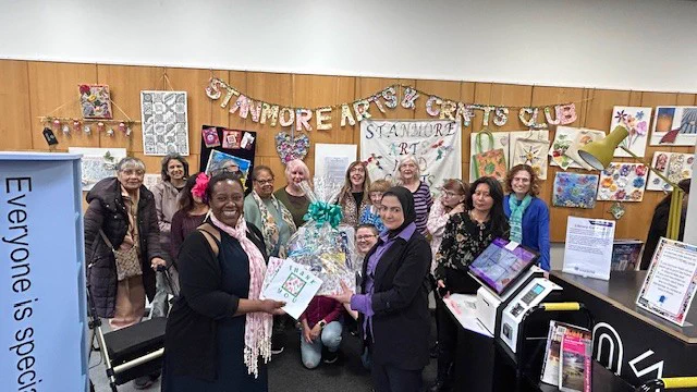 Group of people gathered indoors at Stanmore Arts & Crafts Club, with art displays and a gift basket in the centre.