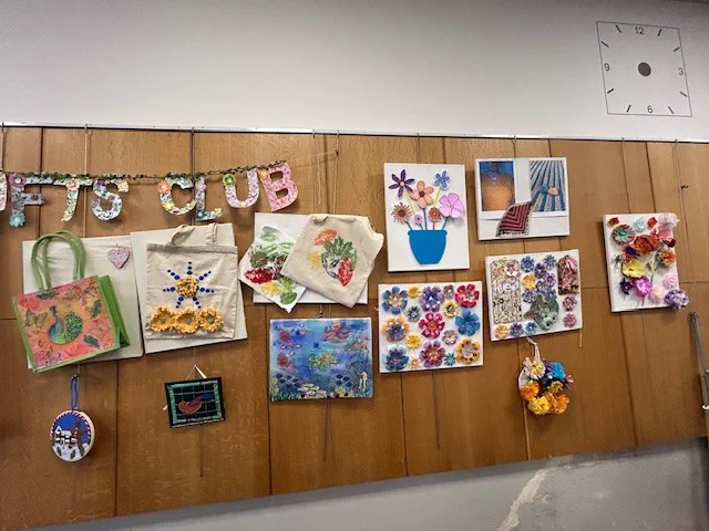 Wall display of colourful handmade crafts and artwork including decorated bags, flower designs, and framed pictures under a floral "ARTS CLUB" sign.