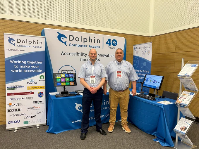 Two men from Dolphin Computer access at the stand at SightCity, the tablet cloths are blue with the Dolphin logo on. On the table are a variety of Dolphin products such as a monitor showing GuideConnect.