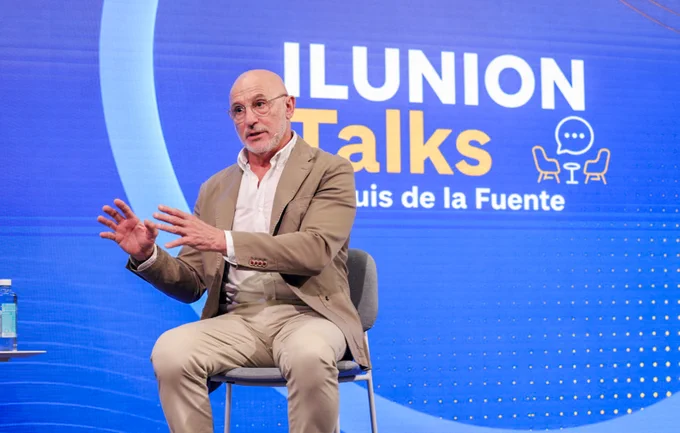 **Texto alternativo:**
Hombre sentado en un escenario durante una charla de ILUNION Talks. Viste chaqueta beige y camisa blanca, lleva micrófono de diadema y gesticula con las manos mientras habla. Al fondo aparece una pantalla azul con el texto “ILUNION Talks” y el nombre “Luis de la Fuente”.
