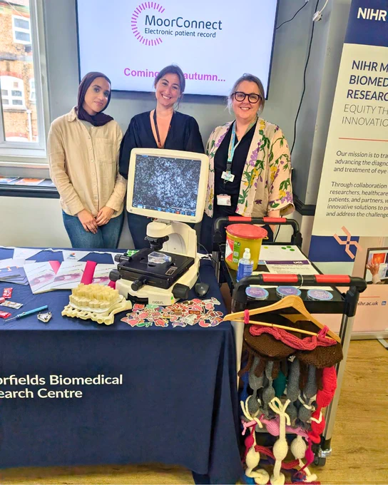 Three people standing behind a research display table featuring a microscope and educational materials. A screen behind them reads “MoorConnect electronic patient record – Coming autumn.” A banner for the NIHR Moorfields Biomedical Research Centre is visible to the side.
