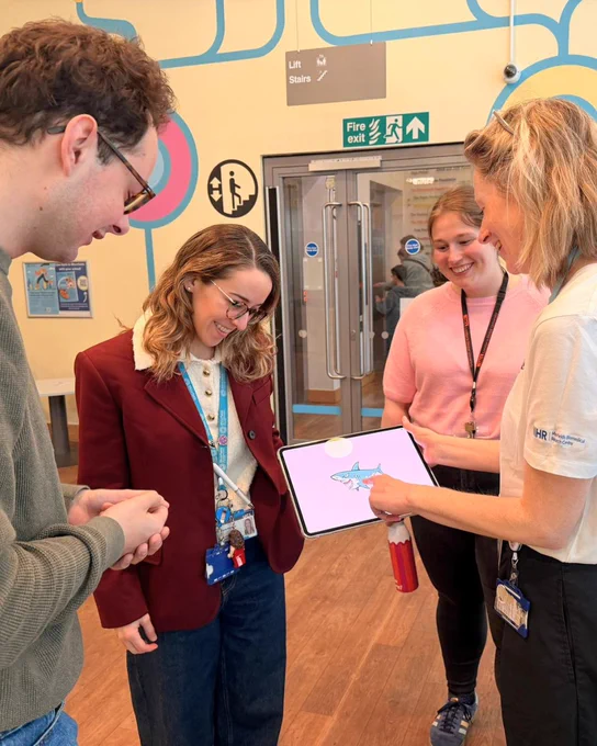 Four people gathered in a hospital corridor around a tablet, which displays a simple illustration of a fish. One person points at the screen while the others look on. Background signage includes directions for lifts, stairs, and a fire exit.
