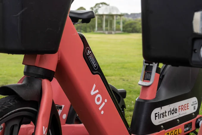 Two red Voi electric scooters parked in a park, with a bandstand in the background. 'voi.' logo visible.