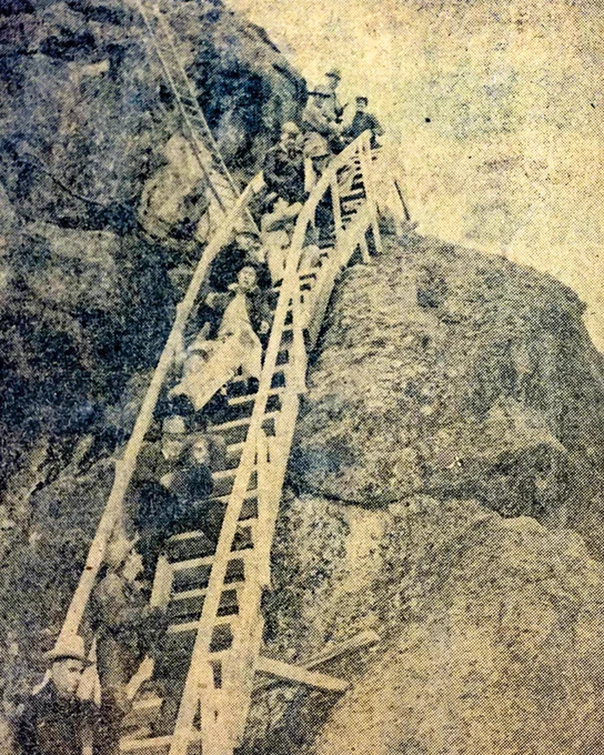 A group of people is seen descending a steep wooden staircase attached to a rocky cliff. The setting appears challenging, with the staircase winding down the rugged terrain. The photograph has an old, grainy quality.