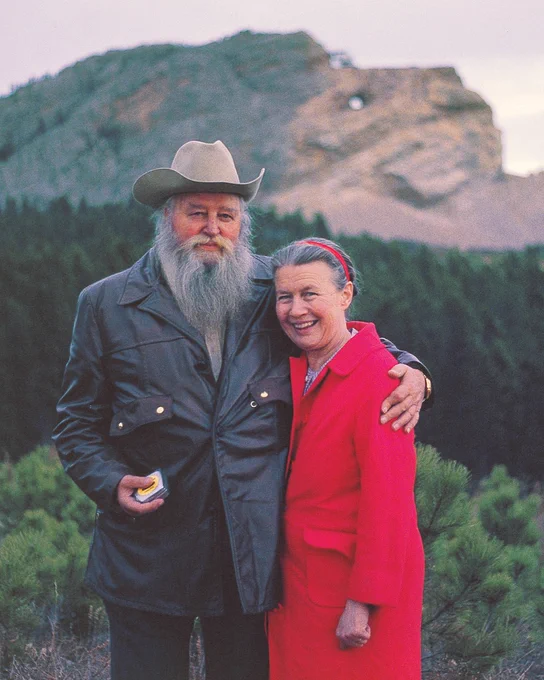 A person wearing a cowboy hat and leather jacket stands beside another individual in a bright red coat. They are outdoors in a natural setting, with trees and the Crazy Horse Memorial visible in the background.