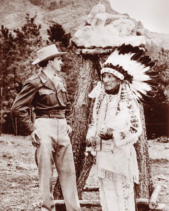 A person in a military uniform stands next to another wearing a feathered headdress and traditional attire. They are outdoors with trees in the background.