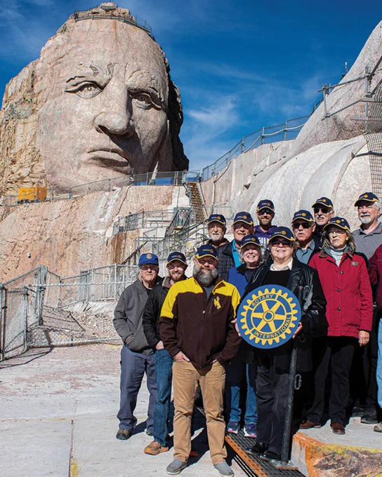 A group of people wearing caps stand in front of the Crazy Horse Memorial. One person holds a Rotary International emblem. The sky is clear and blue.