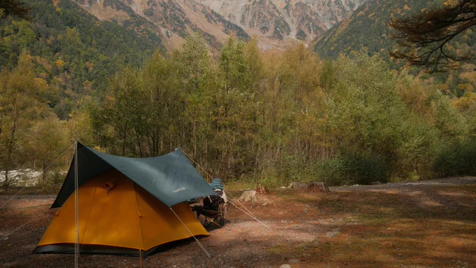 A yellow tent with a dark tarp is set up in a forested area, surrounded by trees with autumn leaves. In the background, there are mountains with sparse vegetation. The scene suggests a peaceful camping setting.