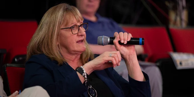 A person is seated in a red auditorium chair, talking into a microphone. 