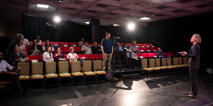 A professional stands on stage speaking to a small audience seated in a theater with red and yellow chairs under bright lights.
