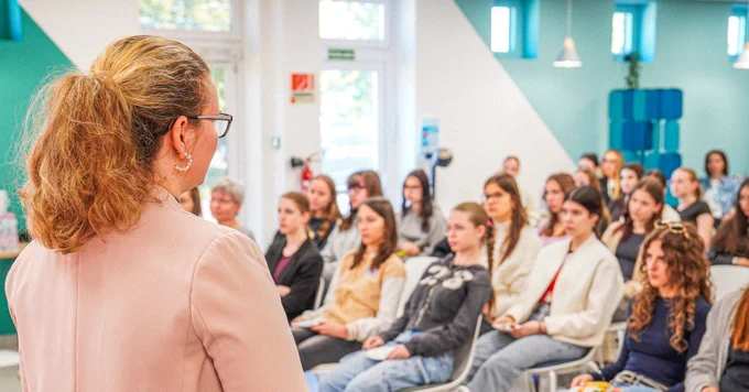 A woman is speaking in front of a seated audience in an indoor setting.