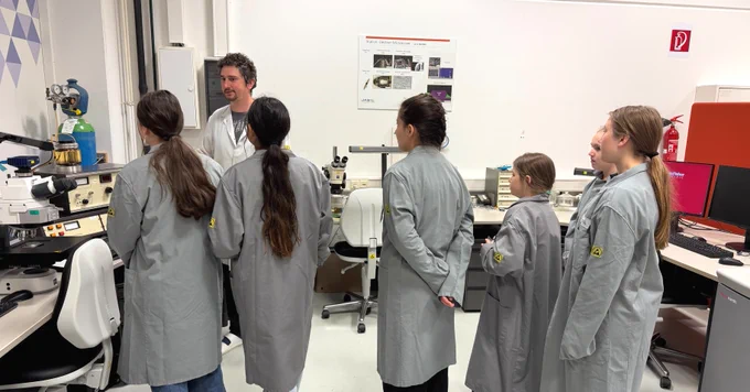 A group of people wearing lab coats are gathered in a laboratory. One person appears to be explaining something. The room contains scientific equipment and posters.