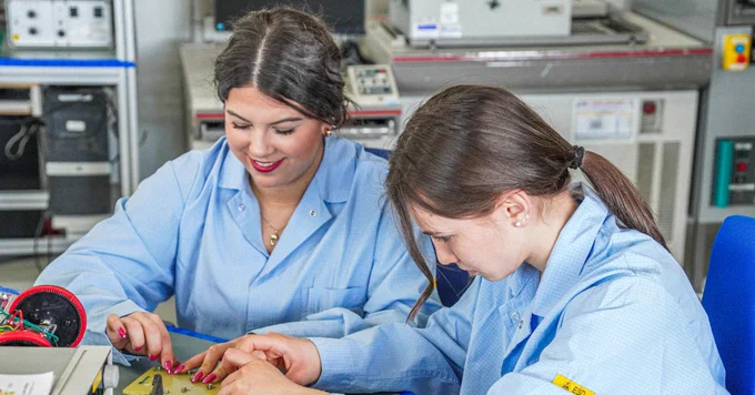 Two people in lab coats work together on a project at a table, surrounded by lab equipment.