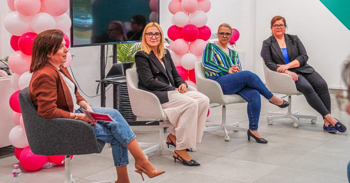 A group of four individuals are seated in front of a room, participating in a panel discussion. They are surrounded by decorative pink and white balloons. A large monitor and some green plants are visible in the background.