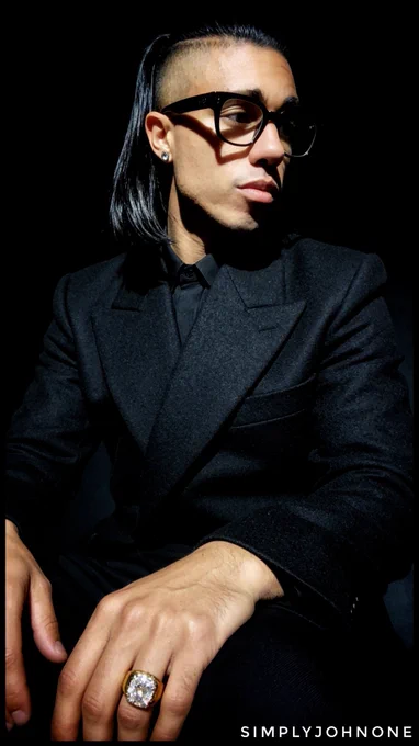 Editorial portrait of a person in a black suit with a gold ring against a dark background.