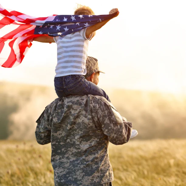 Child sitting on soldier's shoulders waving an American flag in a sunlit field at sunset.