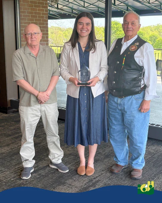 Three people standing indoors near a large window, the woman in the center holds a clear award plaque.