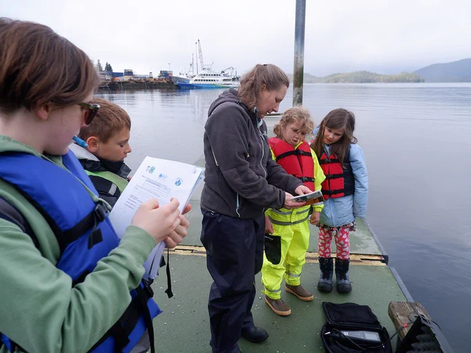 Youths wearing life vests stand on a dock as they fill out forms while an adult explains the process
