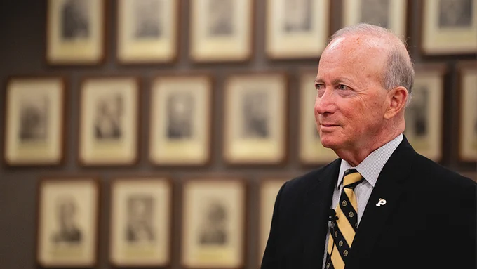 Mitch Daniels wearing a suit with a Purdue University pin. The background features a wall filled with framed sepia-toned portraits.