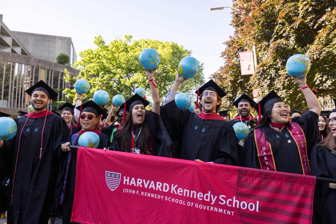 Graduates cheering and holding a Harvard Kennedy School banner and inflatable globes as the process to the celebration.