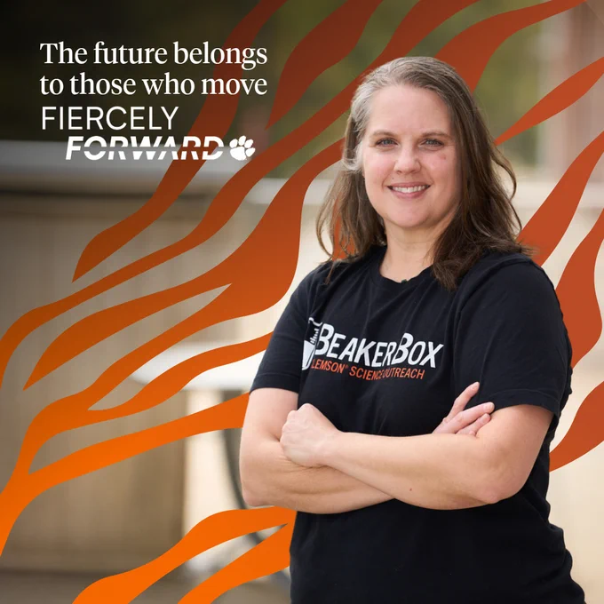 A person smiling while standing with crossed arms. They are wearing a black BeakerBox T-shirt with the text "Clemson Science Outreach." The background features a tiger stripe graphic with the words, "The future belongs to those who move Fiercely Forward."