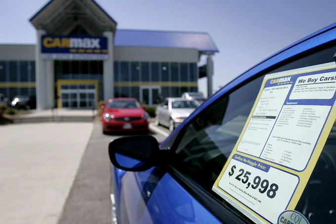 The image shows a CarMax dealership with cars parked outside. A close-up of a blue car's window sticker displays a price of $25,998. The CarMax building has a distinctive blue and yellow sign.