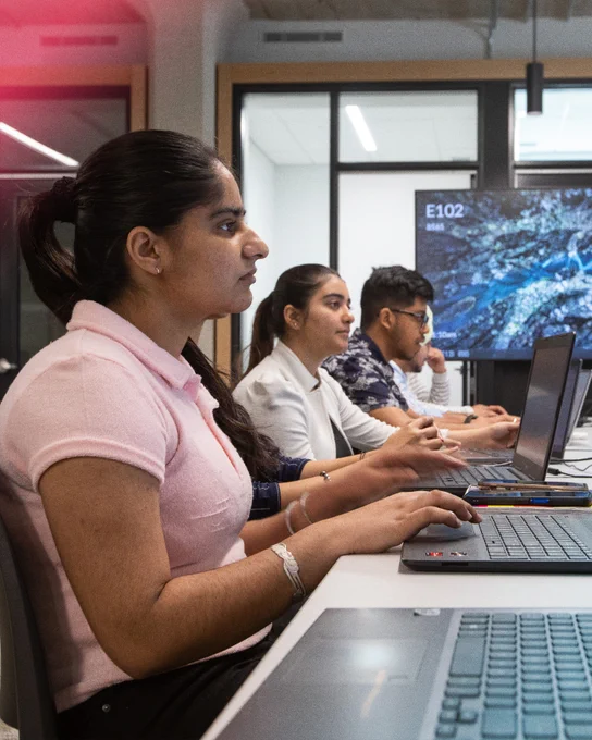 Alt text: Students work on laptops in a classroom, focused on coding and software development activities. Several learners sit side by side at a long table, collaborating and following along with lessons on their computer screens.